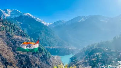 An Indian flag catches the light above a forested mountain valley and distant snowy ridges.