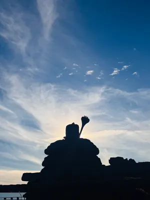 A dark silhouette of a Shiva statue and surrounding stone against a sunlit, cloud-streaked sky.