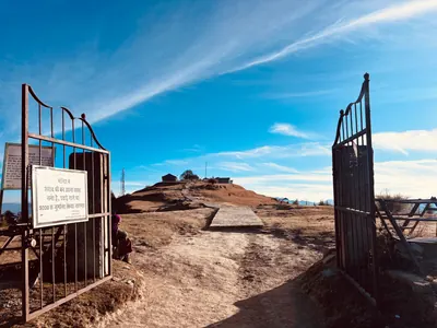 Open iron gates frame a path across a dry hill toward small buildings beneath a blue sky.