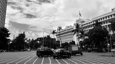 Monochrome street photograph of vehicles, a patterned road, palms and a large pale building.