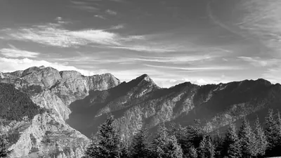 Black-and-white mountain cliffs and forested ridges with conifers in the foreground and streaked clouds above.