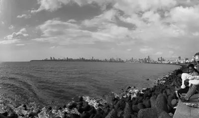 A black-and-white waterfront scene with waves, shoreline blocks, seated visitors and a distant city skyline.