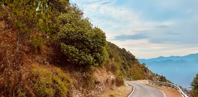 A winding mountain road curves past a wooded hillside toward distant blue ridges.